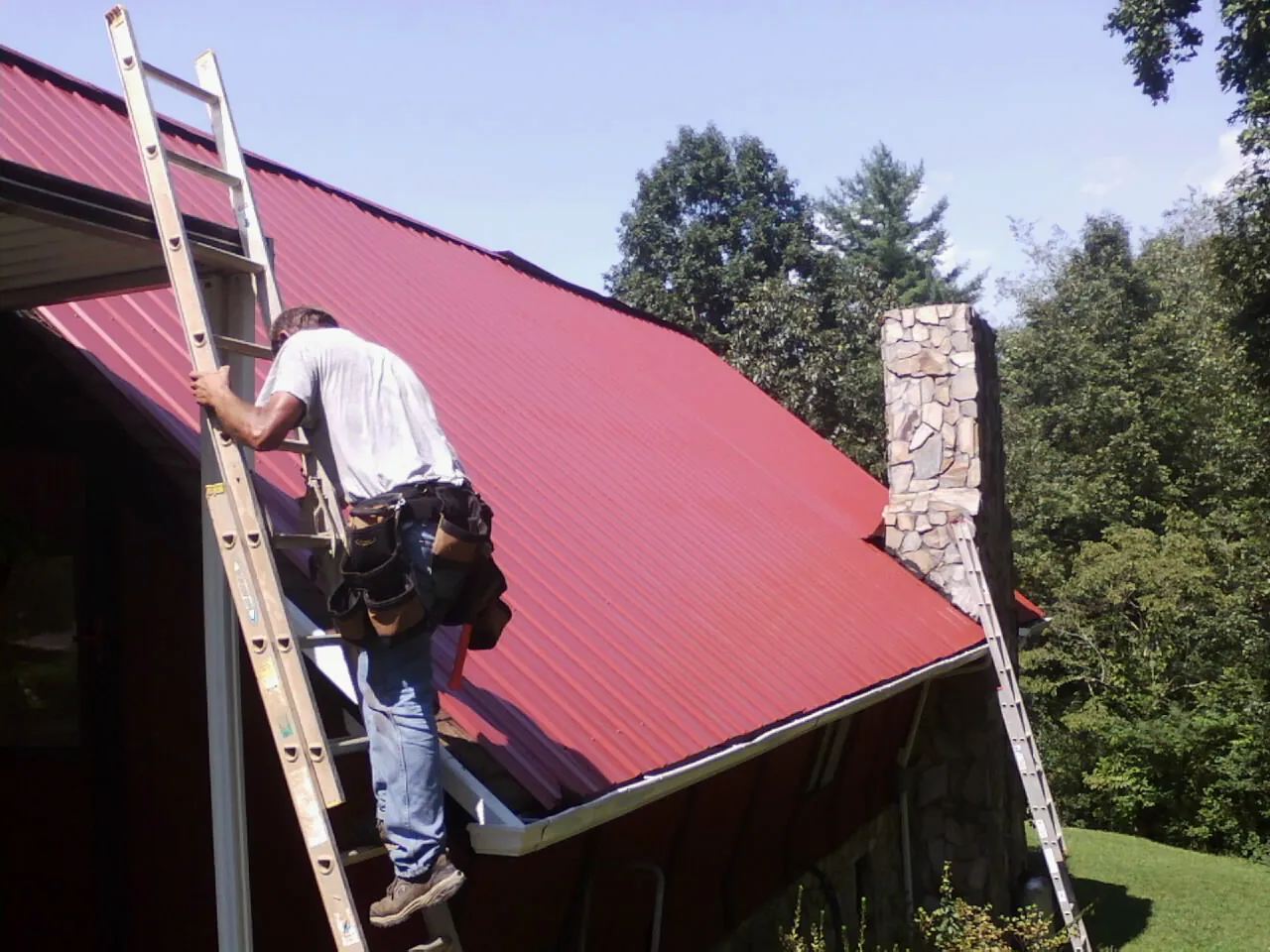 Roofer on a ladder beside a red metal roof and stone chimney — Soffit & Fascia Repair by Roofing Brothers in Pontiac